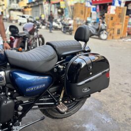 Close-up of a dark blue Royal Enfield Meteor 350 motorcycle with a padded seat and backrest, parked on a busy street storefronts in the background.