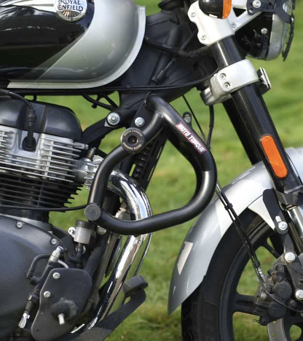 Close-up of a Royal Enfield motorcycle showing the engine, black exhaust pipe, and front fork with an orange reflector on a green background.