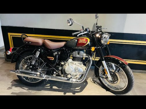 IMG_9711 - laxmiautocare.com Royal Enfield classic motorcycle with brown seat, black fuel tank, and chrome exhaust parked indoors against a black-and-gold wall.