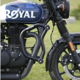 Blue Royal Enfield motorcycle close-up showing the tank with 'ROYAL ENFIELD' branding and exposed engine, crash bars, and front wheel on grass.