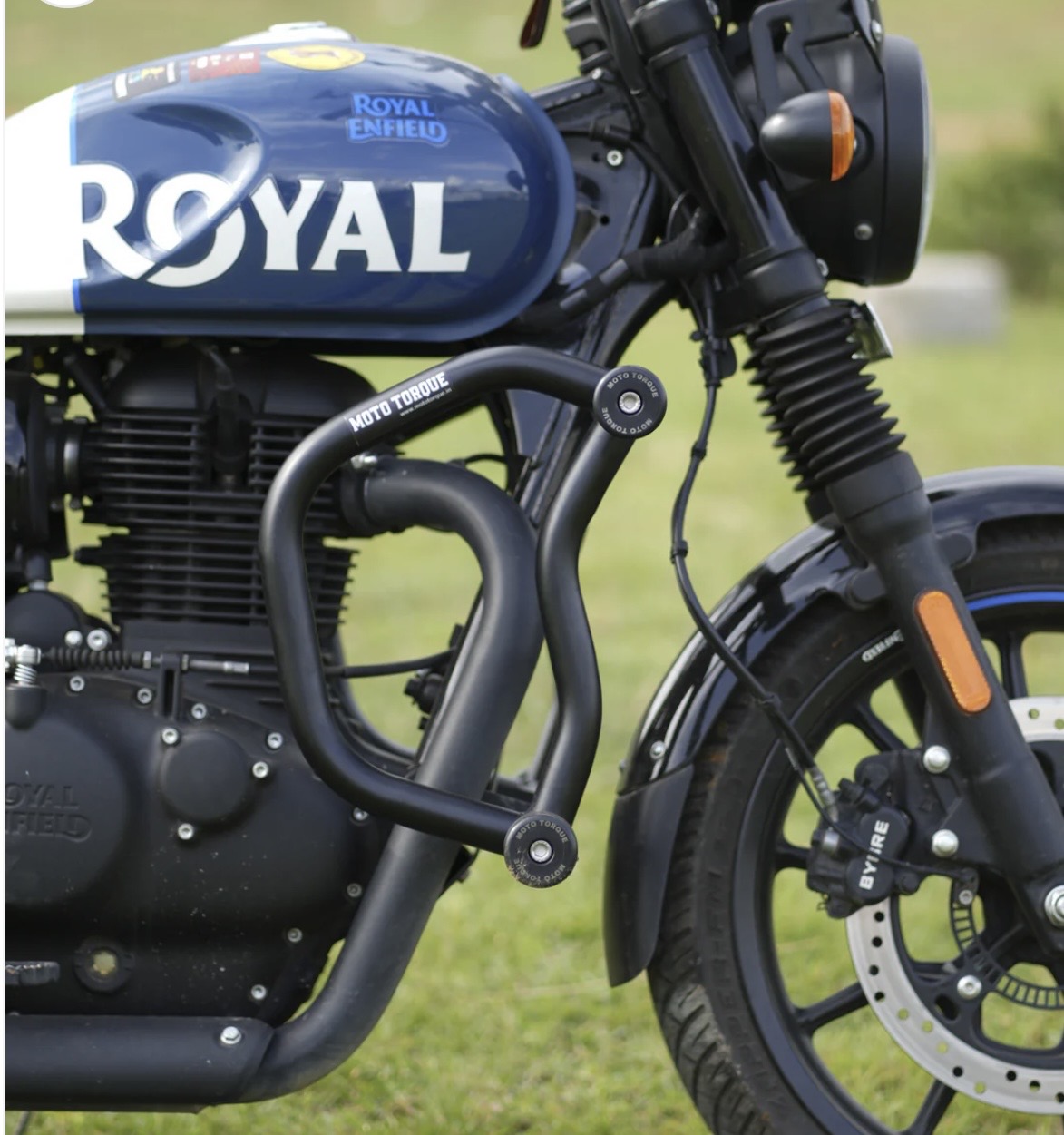 Screenshot - laxmiautocare.com Blue Royal Enfield motorcycle close-up showing the tank with 'ROYAL ENFIELD' branding and exposed engine, crash bars, and front wheel on grass.