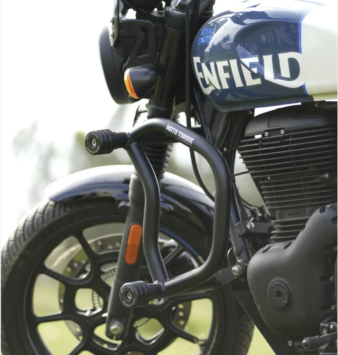 Screenshot - laxmiautocare.com Close-up of a Royal Enfield motorcycle front, showing crash bars with 'Moto Torque', front wheel, and blue-and-white fuel tank.