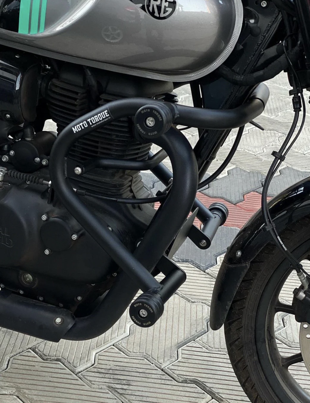 Screenshot - laxmiautocare.com Close-up of a matte-black motorcycle engine and frame with a curved crash bar and a 'Moto Torque' pipe/branding visible on the tube near the tank edge.
