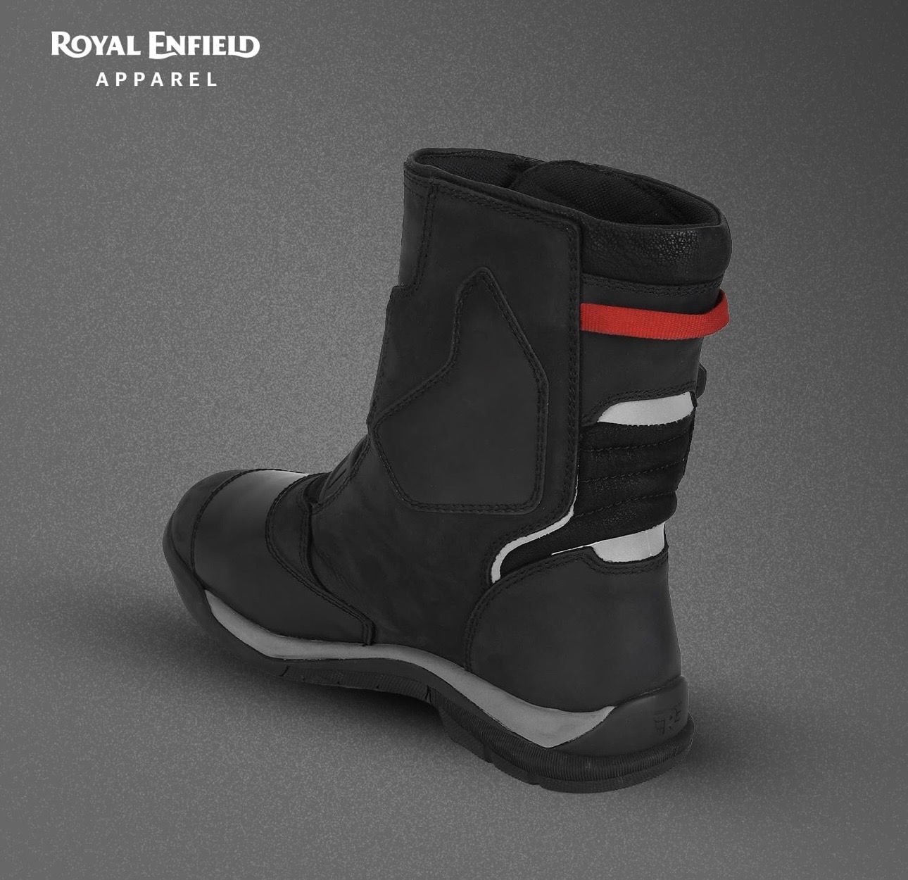 Screenshot - laxmiautocare.com Black motorcycle boot shown from a rear-side angle on a gray studio background; features a red strap and white accents, with Royal Enfield Apparel branding in the top-left corner