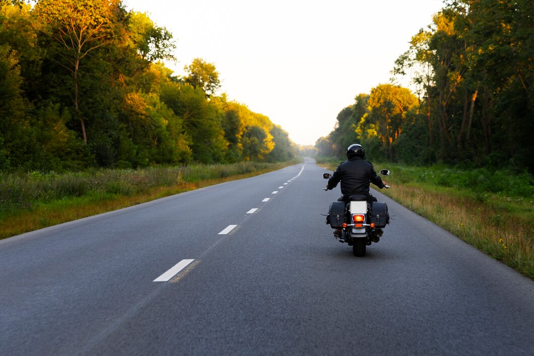 Motorcycle rider in black gear travels down a rural two-lane road lined with trees at sunset-like light or golden hour.