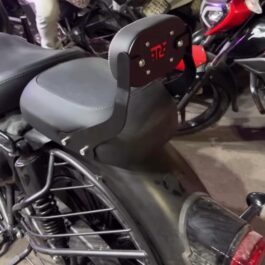 85ed1077-c7e9-4945-85c9-ff6aba57cb63 - laxmiautocare.com Motorcycle with a black backrest mounted behind the seat, featuring a red logo and silver bolts, parked among other bikes indoors.