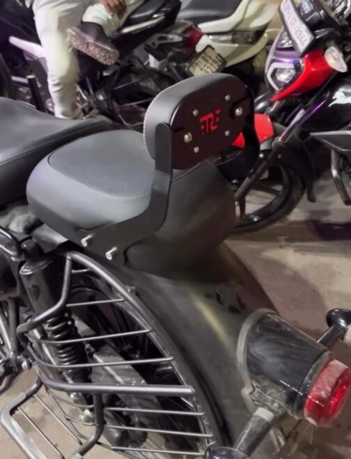 85ed1077-c7e9-4945-85c9-ff6aba57cb63 - laxmiautocare.com Motorcycle with a black backrest mounted behind the seat, featuring a red logo and silver bolts, parked among other bikes indoors.