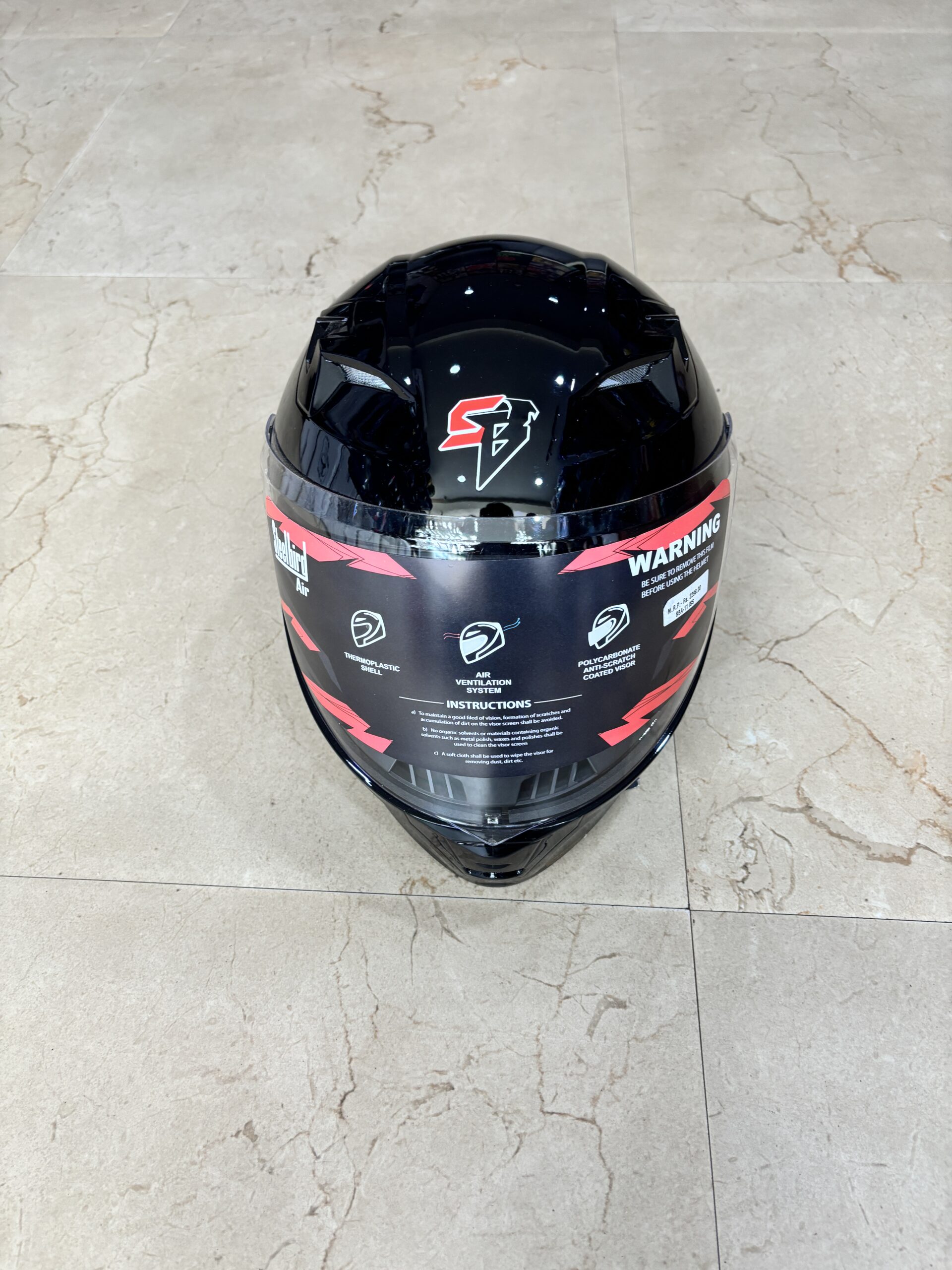 IMG_2180 - laxmiautocare.com Top view of a glossy black full-face motorcycle helmet on a light beige tiled floor, with a red warning label around the rim.