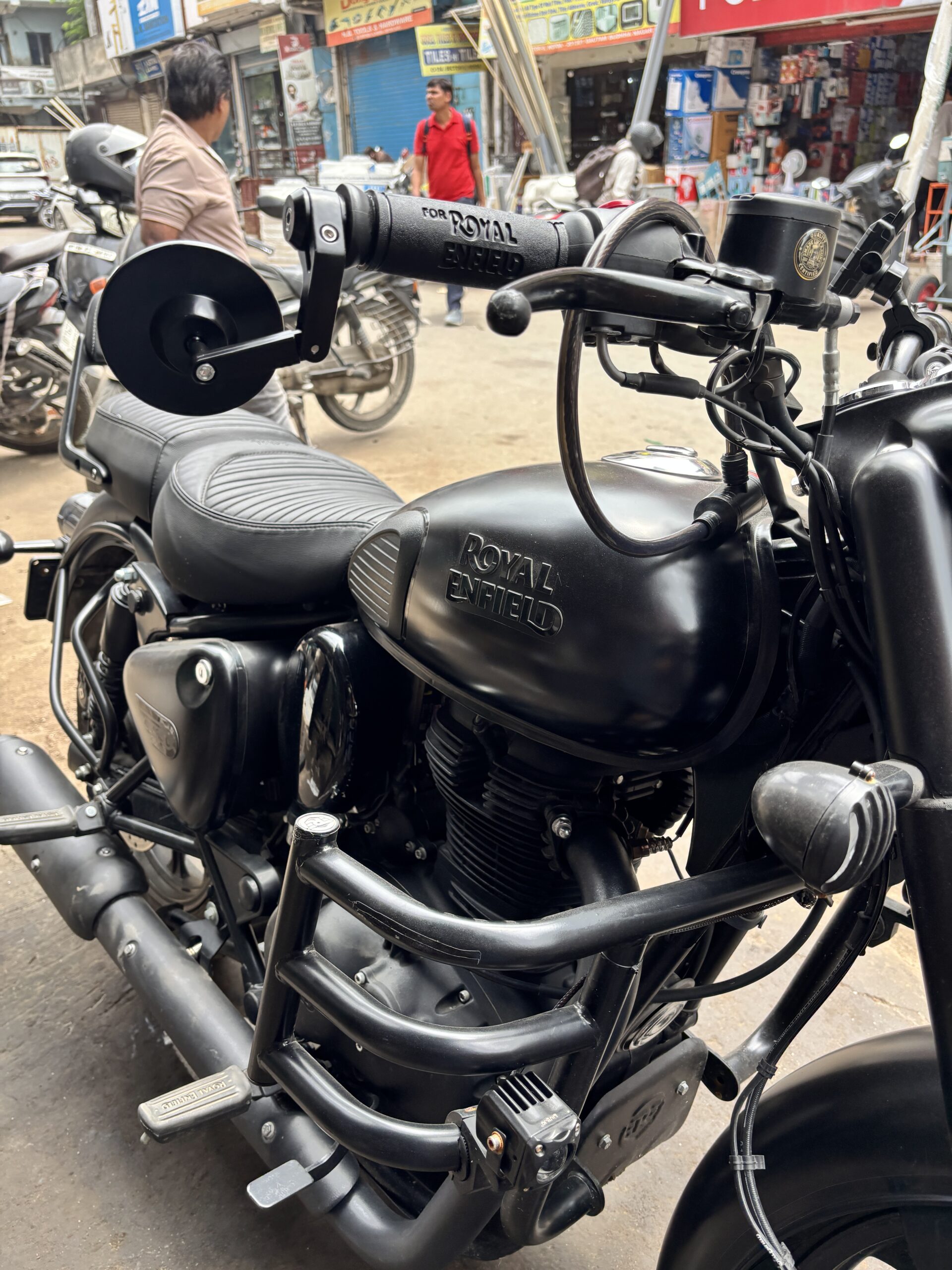 IMG_3391 - laxmiautocare.com Close-up of a black Royal Enfield motorcycle showing the handlebars, mirror mount, and the fuel tank with branding, with a busy street market in the background.