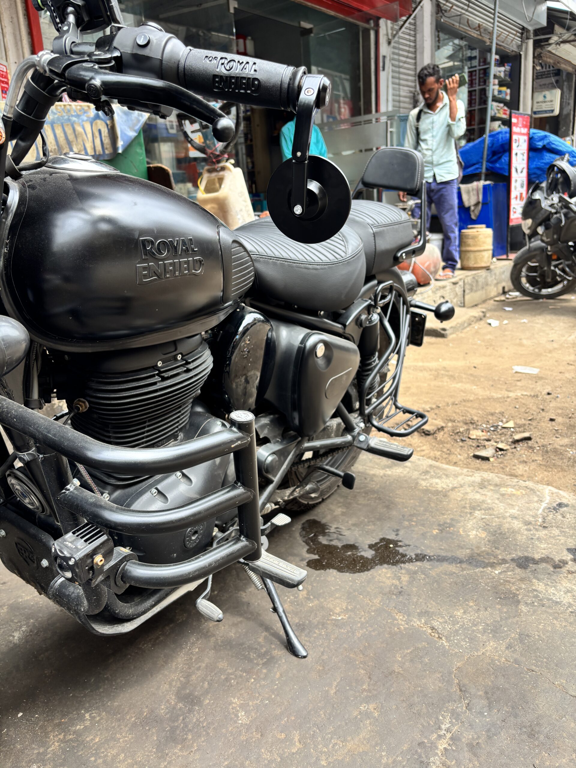 IMG_3392 - laxmiautocare.com Close-up of a black Royal Enfield motorcycle on a street, showing the seat and handlebars with a man in a light shirt in the background.