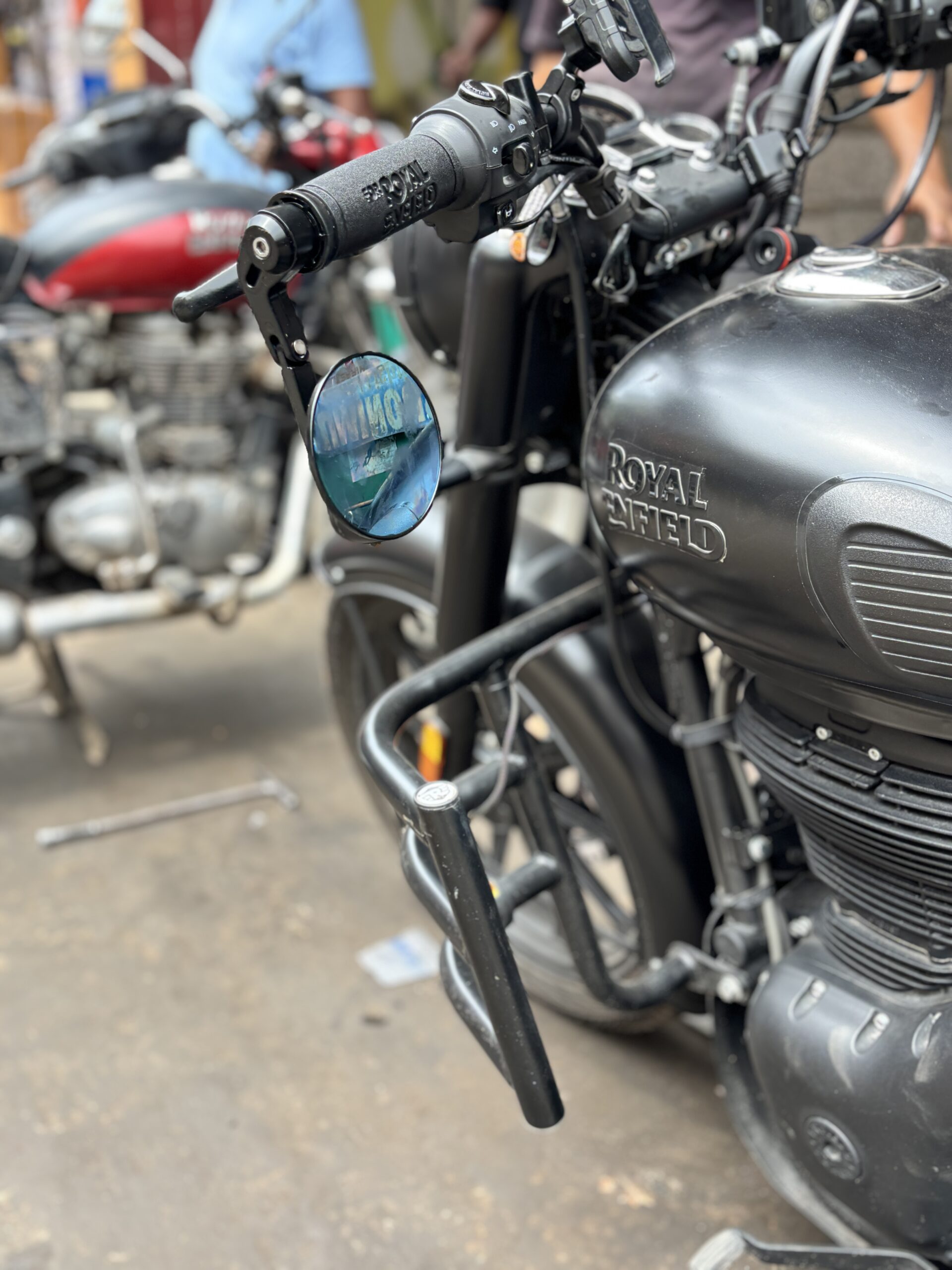 IMG_3393 - laxmiautocare.com Close-up of a black Royal Enfield motorcycle handlebar and mirror, with the blue-tinted round mirror reflecting a street scene.