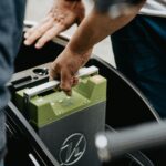 Close-up of hands handling a green car battery in the trunk of a vehicle, preparing to install or remove it.