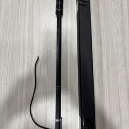 Black cylindrical telescoping tool with a red tip, silver ring accents, placed beside a tall black rectangular case on a light wood surface. A black cord runs near the bottom left.