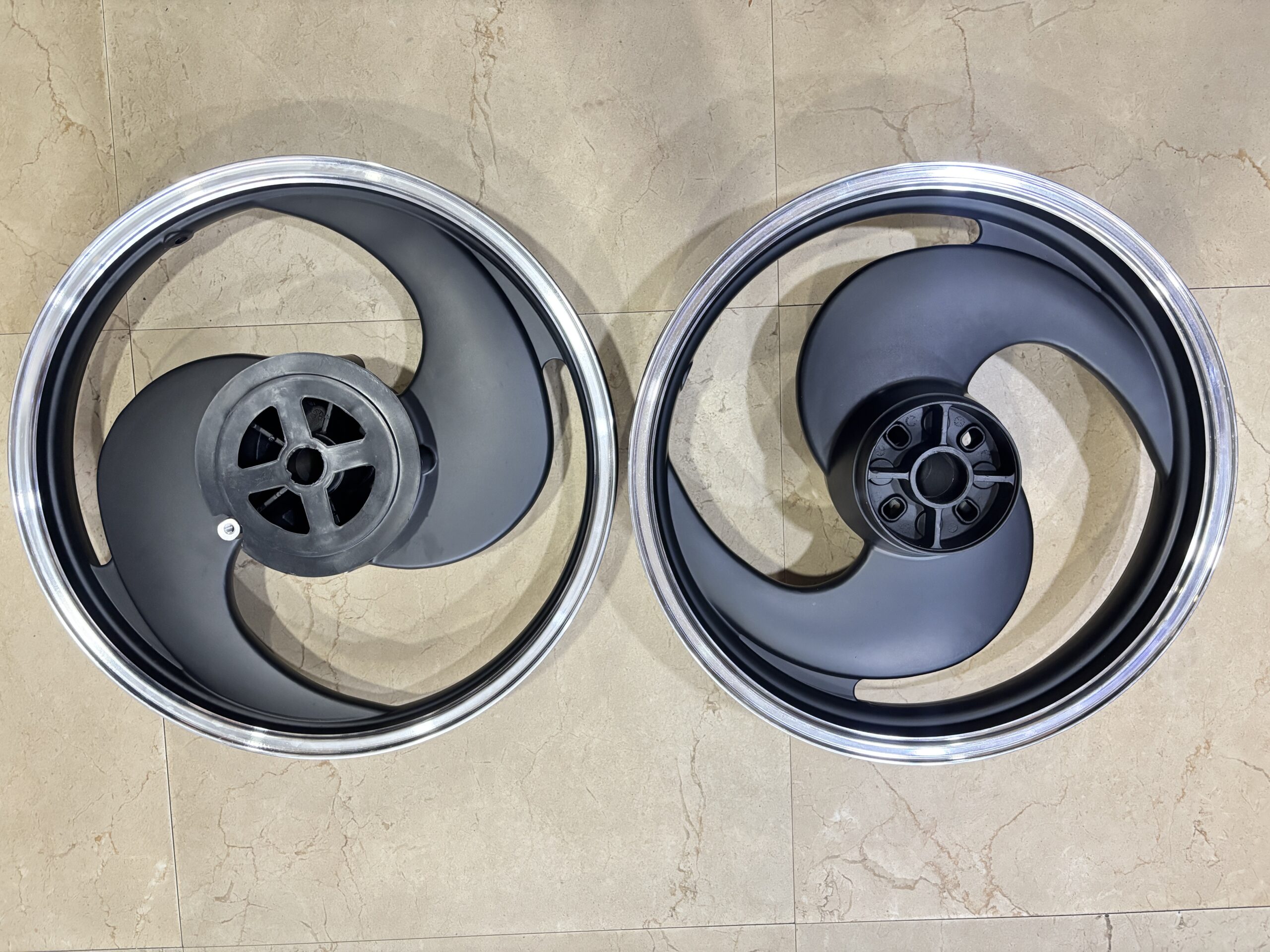IMG_5705 - laxmiautocare.com Two alloy car wheels lying flat on a beige tiled floor, showing dark gray spokes and shiny silver rims.