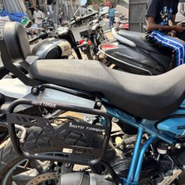 Close-up of a blue motorcycle seat and metal rack, with other bikes and a busy market street in the background.