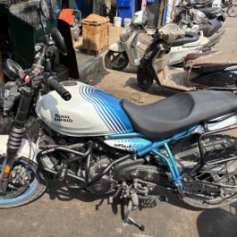 Blue and white Royal Enfield motorcycle in the foreground among other scooters parked on a busy street outside a shop.