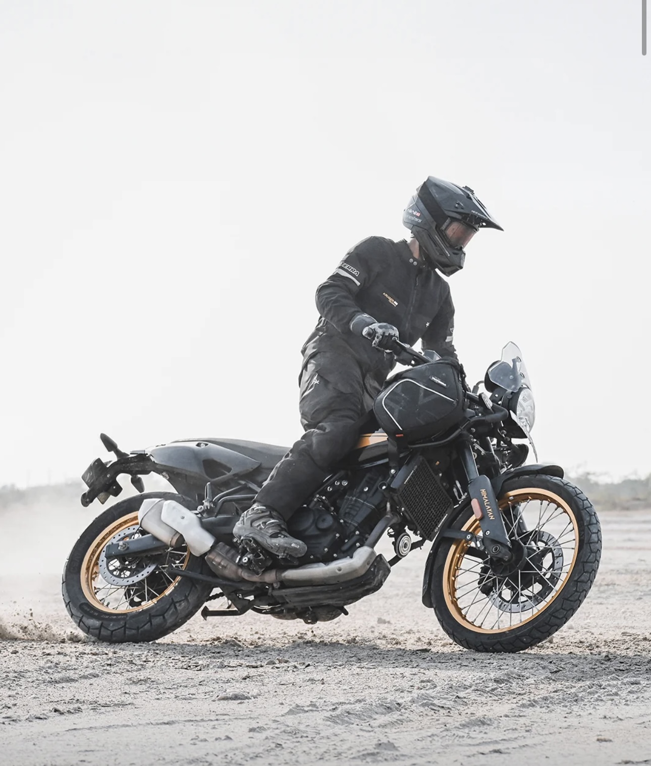 Screenshot - laxmiautocare.com Motorcyclist in protective gear sitting astride a black motorcycle with gold rims on a dusty off-road trail