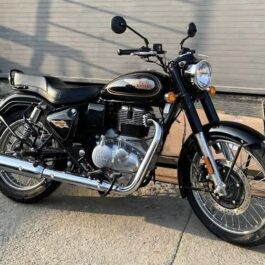 Classic black retro motorcycle with chrome exhaust and round headlight parked beside a roller shutter; single long seat and mirrors visible.